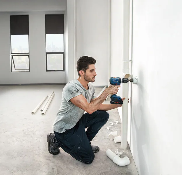 Una persona perfora una pared para instalar tuberías en una habitación luminosa y sin terminar.