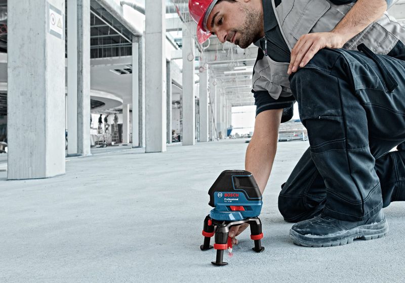 Eine Person mit Sicherheitsausrüstung richtet ein Lasernivelliergerät auf einem Betonboden aus.