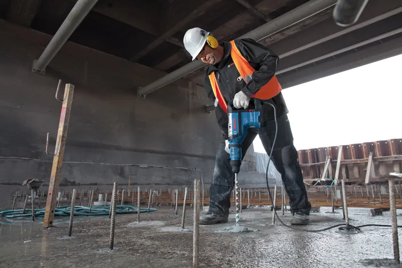 Eine Person mit Sicherheitsausrüstung bohrt auf einer Baustelle mit einem Bohrhammer in Beton.
