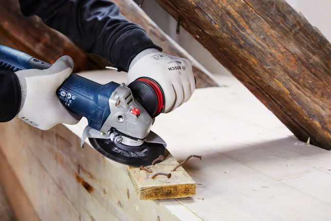 Ein Arbeiter mit Sicherheitsausrüstung schneidet mit einem Winkelschleifer Nägel in ein Holzbrett.