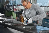 Eine Person mit Schutzausrüstung schleift mit einem Winkelschleifer Metallträger, wobei Funken entstehen.