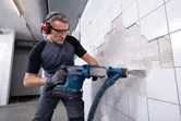 Eine Person mit Schutzausrüstung entfernt Wandfliesen mit einem Akku-Bohrhammer.