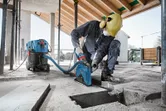 Eine Person mit Sicherheitsausrüstung schneidet eine Nut in Beton, angeschlossen an einen Nass-/Trockensauger.