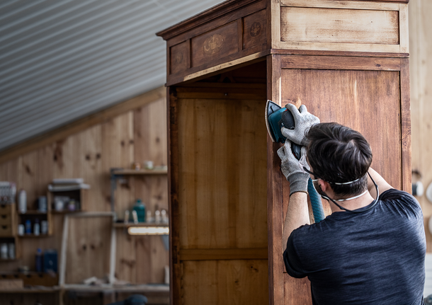 Person mit Sicherheitsausrüstung schleift in einer Werkstatt einen Holzschrank.