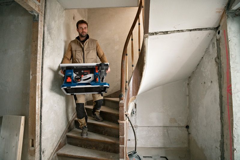 Eine Person mit Sicherheitsausrüstung trägt eine Akku-Tischkreissäge eine Treppe hinunter.