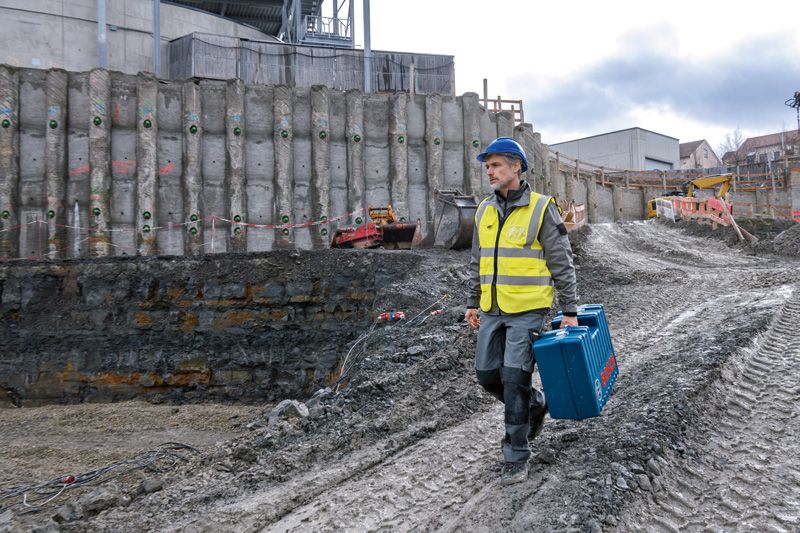 Eine Person mit Sicherheitsausrüstung trägt einen blauen Werkzeugkoffer auf einer schlammigen Baustelle.