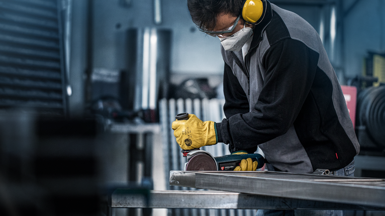 Eine Person, die Schutzausrüstung trägt, schleift einen Metallträger mit einem Elektrowerkzeug.
