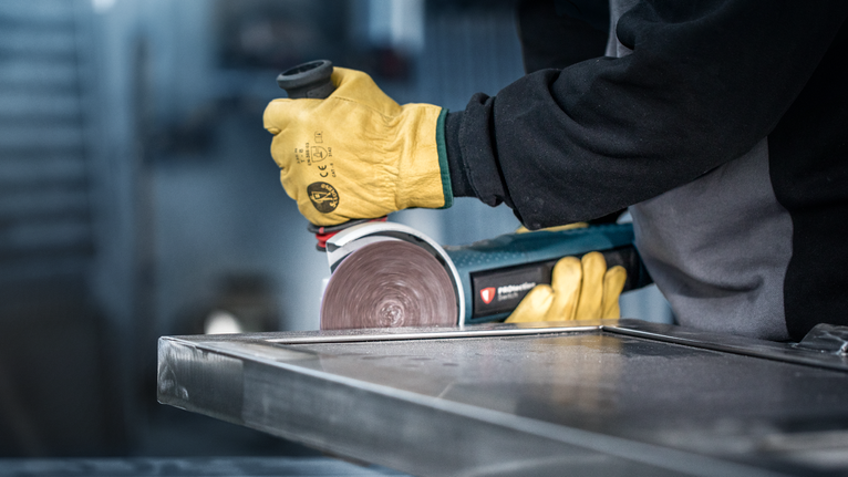 Eine Person, die Schutzausrüstung trägt, schleift ein Metallblech mit einem Winkelschleifer.