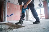 Eine Person mit Sicherheitsausrüstung verwendet einen Akku-Bohrhammer, um Beton aufzubrechen.