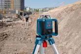Laser-Nivelliergerät auf einem Stativ auf einer Baustelle mit einer Person in Schutzausrüstung.