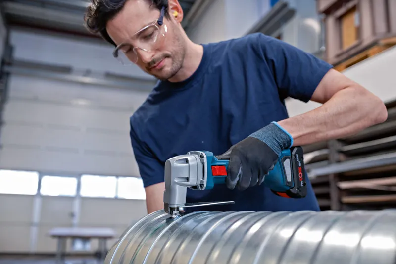 Eine Person mit Sicherheitsausrüstung schneidet mit einer kabellosen Metallschere einen Metallkanal durch.