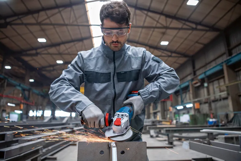 Eine Person, die Schutzausrüstung trägt, schleift in einer Fabrik Metall mit einem Akku-Winkelschleifer.