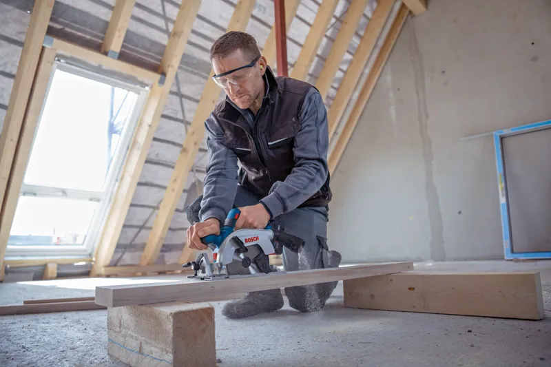 Eine Person mit Sicherheitsausrüstung schneidet in einem unausgebauten Dachboden mit einer Kreissäge ein Holzbrett.