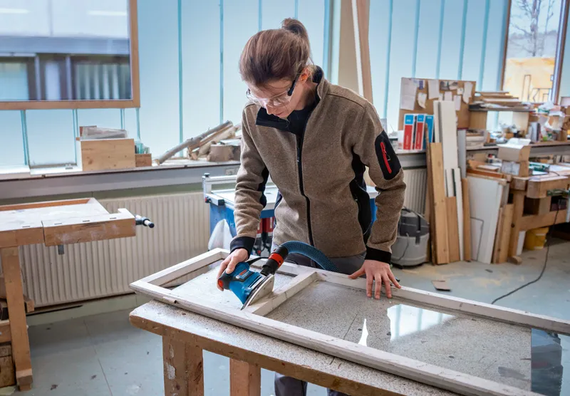 Eine Person mit Schutzausrüstung schleift in einer Werkstatt einen hölzernen Fensterrahmen.
