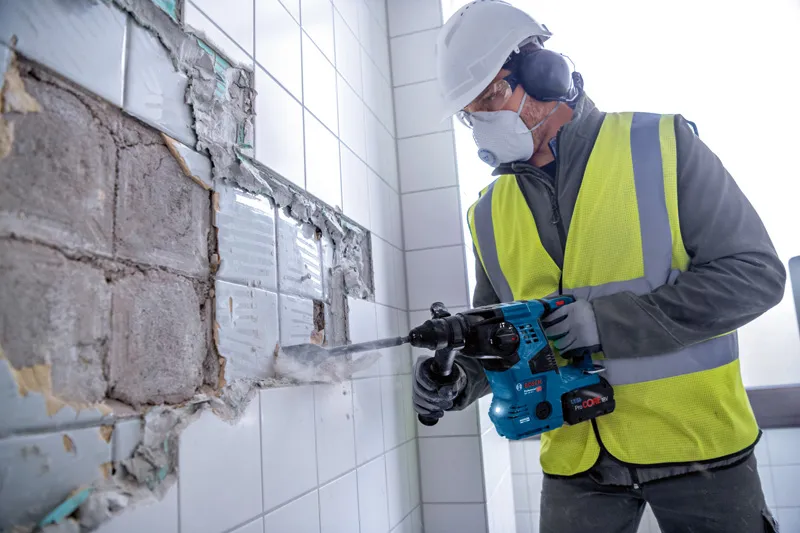 Eine Person mit Schutzausrüstung entfernt mit einem Bohrhammer Fliesen aus einer Wand.