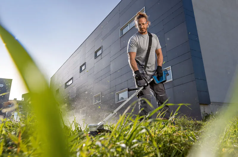 Eine Person mit Schutzausrüstung schneidet hohes Gras mit einem Akku-Rasentrimmer.