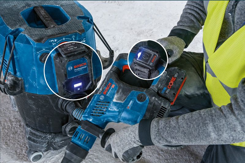 Eine Person, die Schutzausrüstung trägt, benutzt ein Elektrowerkzeug, das an eine Staubabsaugung angeschlossen ist.