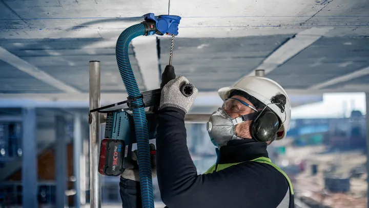 Eine Person mit Sicherheitsausrüstung bohrt auf einer Baustelle in eine Betondecke.