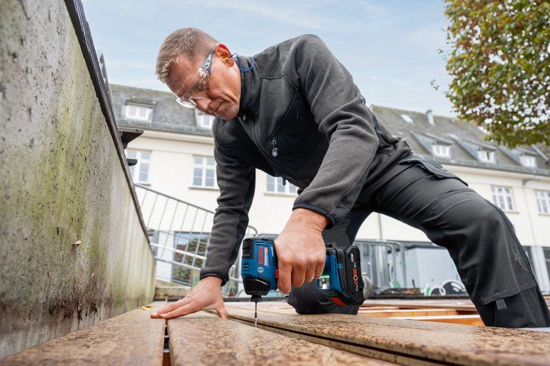 Eine Person mit Sicherheitsausrüstung bohrt in Holzbretter auf einem Außendeck.