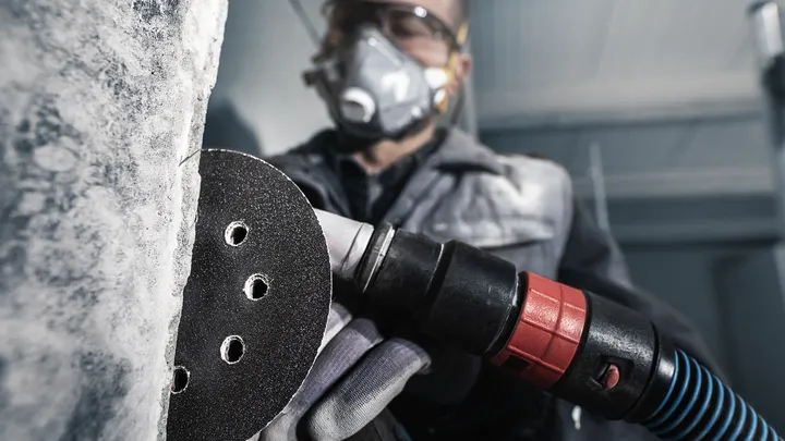 Eine Person mit Sicherheitsausrüstung schleift mit einem Elektrowerkzeug eine Betonwand.