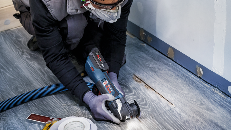 Eine Person mit Sicherheitsausrüstung schneidet mit einem Elektrowerkzeug einen Holzboden.