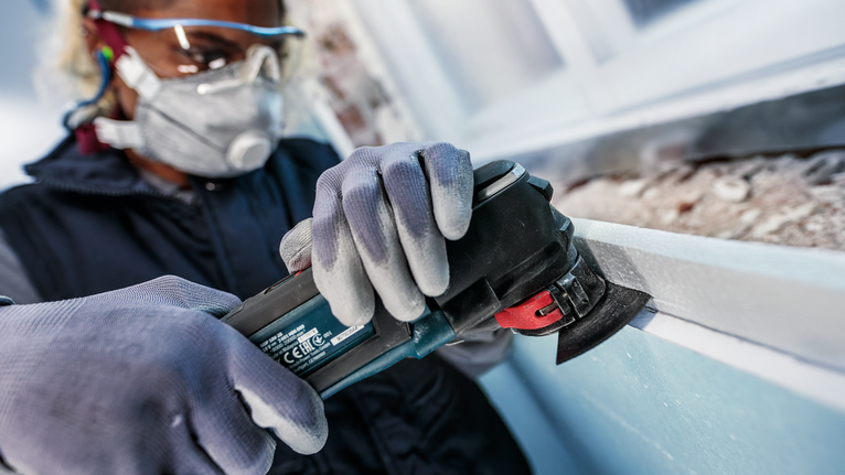 Eine Person mit Schutzausrüstung schleift mit einem Elektrowerkzeug einen Fensterrahmen.