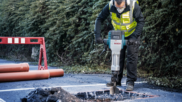 Eine Person mit Sicherheitsausrüstung bricht mit einem Abbruchhammer Asphalt auf einer Straße auf.