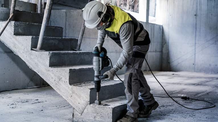 Eine Person mit Sicherheitsausrüstung bohrt in Beton in der Nähe einer Treppe.