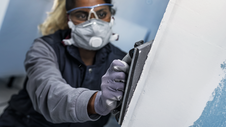Eine Person mit Sicherheitsausrüstung schleift eine Wand mit einem Handschleifer.