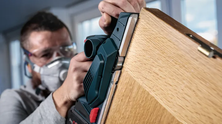 Eine Person mit Schutzausrüstung hobelt die Kante einer Holztür mit einem Elektrowerkzeug.