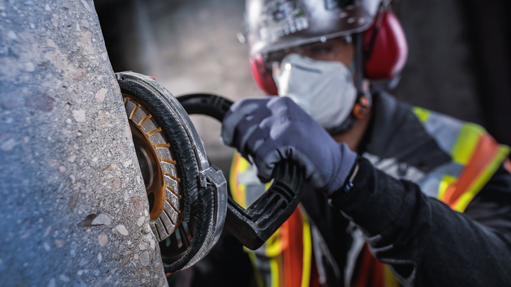 Eine Person mit Sicherheitsausrüstung schleift Beton mit einem Elektrowerkzeug.