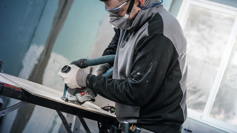 Eine Person mit Sicherheitsausrüstung schneidet Holz mit einem an einen Vakuumschlauch angeschlossenen Elektrowerkzeug.