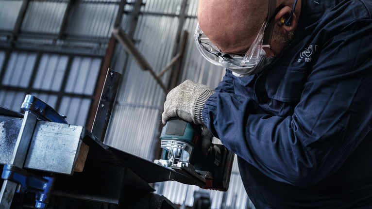 Eine Person mit Schutzausrüstung schleift die Kante eines Metallträgers.