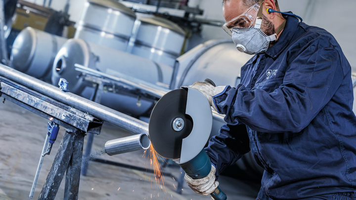 Eine Person mit Sicherheitsausrüstung schneidet mit einem großen Winkelschleifer ein Metallrohr.