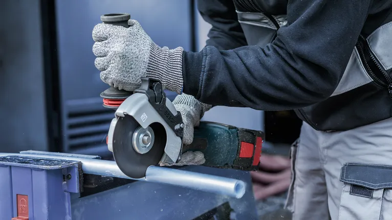 Eine Person mit Sicherheitsausrüstung verwendet einen Winkelschleifer, um ein Metallrohr zu schneiden.