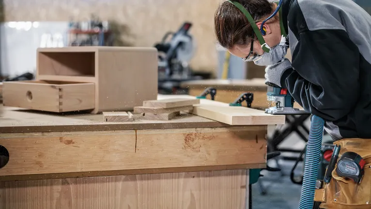 Eine Person mit Sicherheitsausrüstung formt Holzbretter mit einem Elektrowerkzeug auf einer Werkbank.