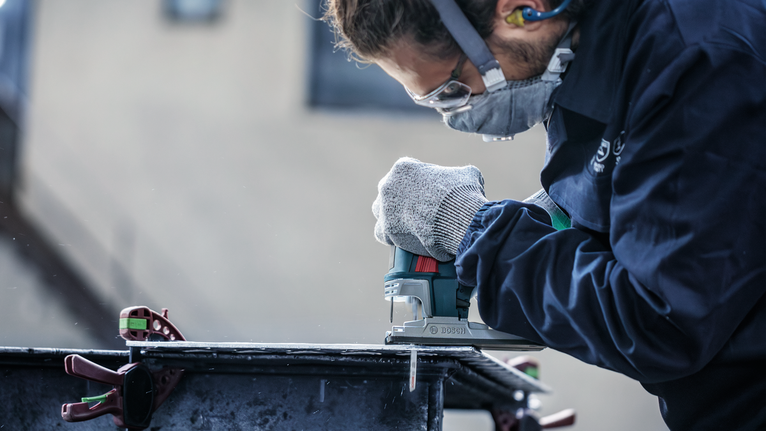 Eine Person mit Sicherheitsausrüstung schneidet mit einer Stichsäge ein Metallblech.