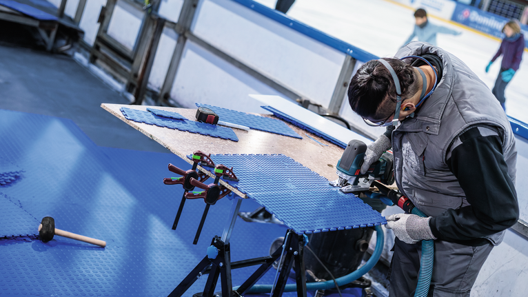 Person mit Schutzausrüstung schneidet blaue Bodenplatten mit einem Elektrowerkzeug.