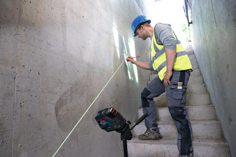 Eine Person mit Sicherheitsausrüstung markiert mit einem Lasernivelliergerät eine Wand auf einer Treppe.