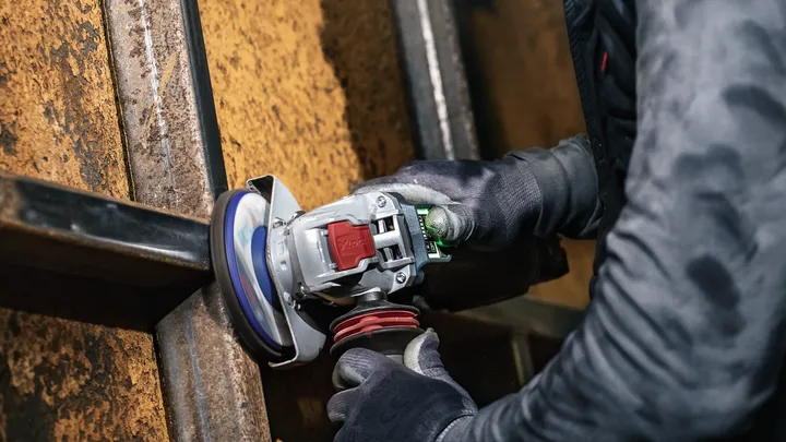 Eine Person mit Schutzausrüstung schleift einen Metallträger mit einem Winkelschleifer.