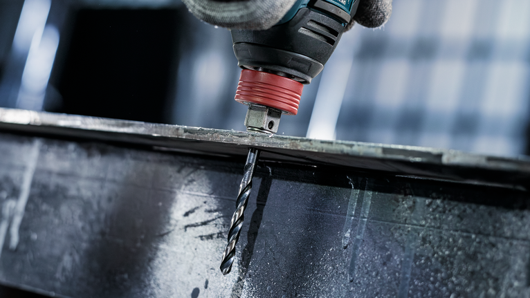 Person mit Sicherheitsausrüstung bohrt durch einen Metallträger.