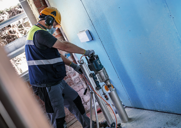 Person mit Schutzausrüstung bohrt mit montiertem Elektrowerkzeug in eine blaue Wand.