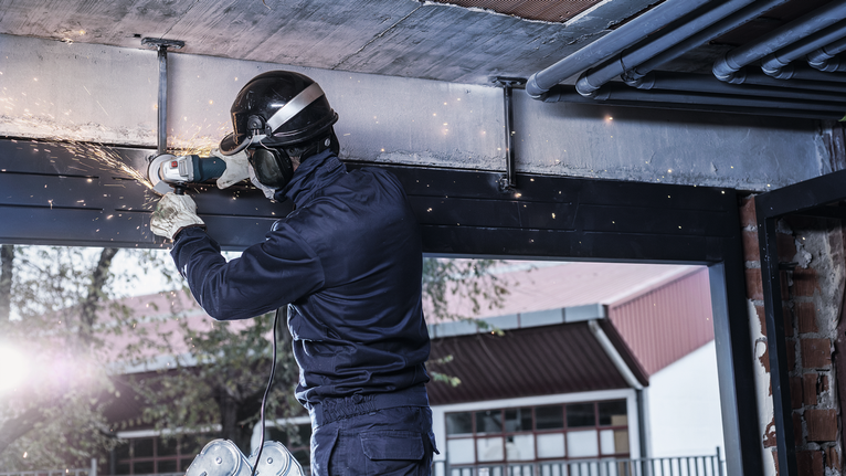Eine Person mit Sicherheitsausrüstung schleift einen Metallträger und erzeugt dadurch im Innenbereich Funken.
