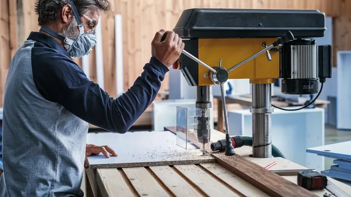 Eine Person mit Sicherheitsausrüstung bedient eine Bohrmaschine, um Löcher in Holz zu bohren.