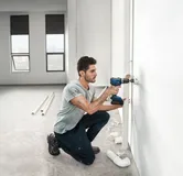 Eine Person bohrt in eine Wand, um in einem hellen, unfertigen Raum Rohrleitungen zu installieren.