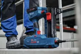 Ein kabelloser Betonnagler liegt auf einer Baustellenfläche in der Nähe einer Person, die Schutzausrüstung trägt.