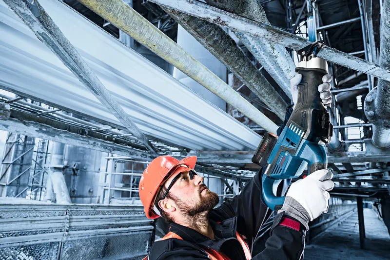 Eine Person mit Sicherheitsausrüstung schneidet über dem Kopf mit einer Säbelsäge ein Metallrohr durch.