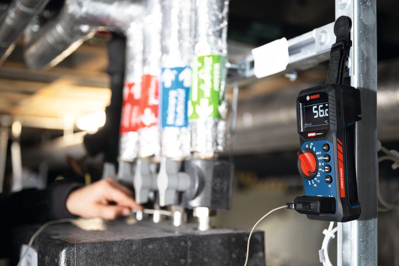 Eine Person inspiziert Rohre, während ein digitales Multimeter in der Nähe die Temperatur misst.