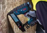 Eine Person, die Schutzausrüstung trägt, benutzt einen kabellosen Tacker mit mittlerer Krone an einer Holzplatte.