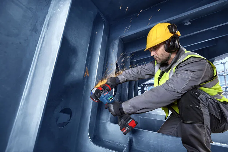 Eine Person mit Sicherheitsausrüstung schleift Metall in einer großen Stahlkonstruktion.